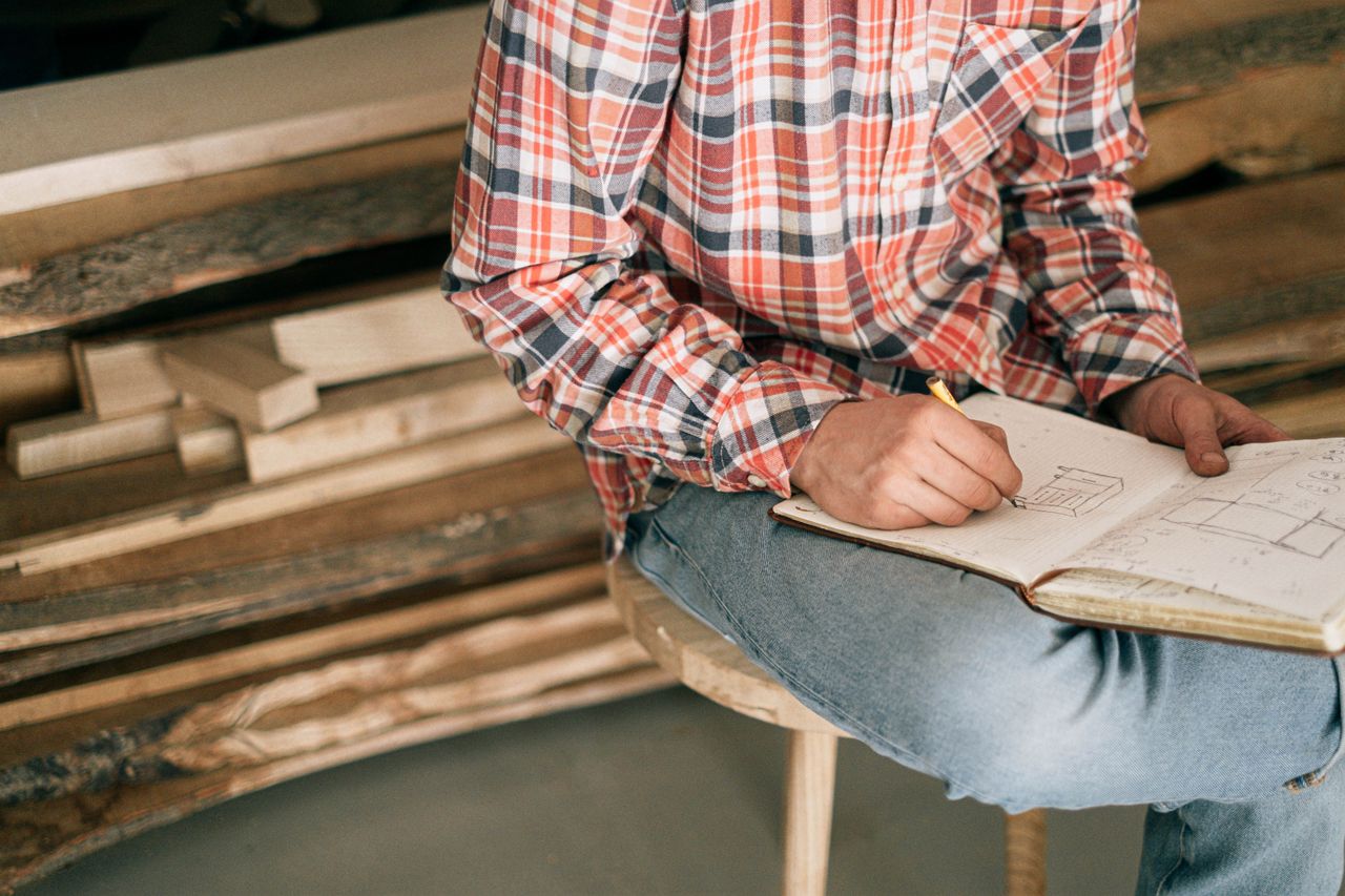 A carpenter in a workshop sketches furniture designs in a notebook, surrounded by wood planks.